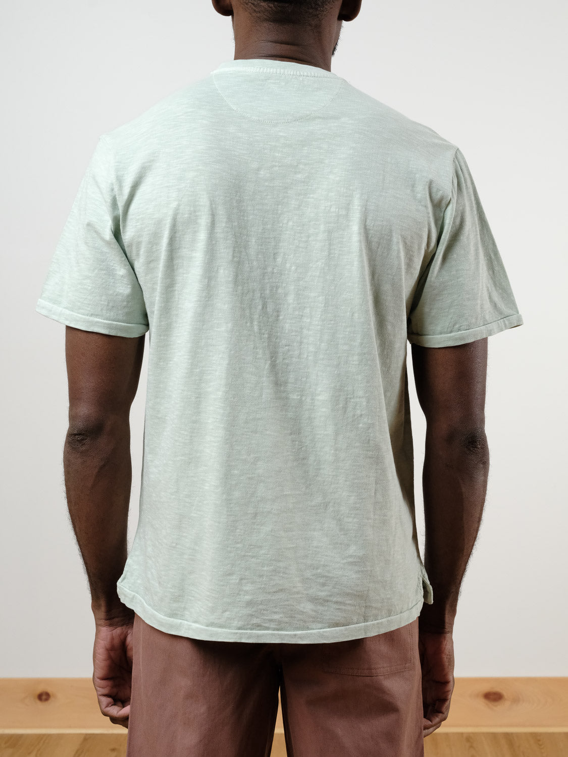 A person stands indoors facing away from the camera, wearing the light green Rogue Henley short-sleeve t-shirt by ID Menswear and brown pants. The setting features a plain white wall and a wooden floor.