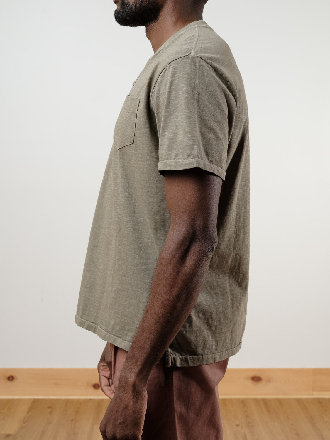 A man stands in profile wearing the Rogue Henley from ID Menswear, a khaki 100% cotton short-sleeve t-shirt with chest pocket, paired with brown pants. His face isn’t visible. The plain background has light walls and a wooden floor.