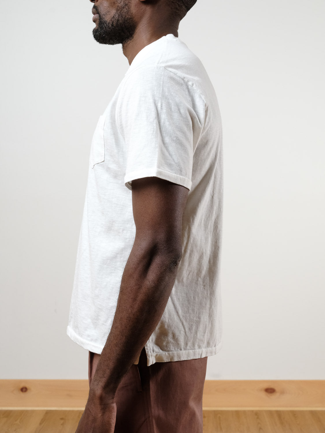 A man in profile against a plain background wears the ID Menswear Rogue Henley—a white, short-sleeve 100% cotton T-shirt with a chest pocket—paired with brown pants. The photo is cropped above his head and below his knees.