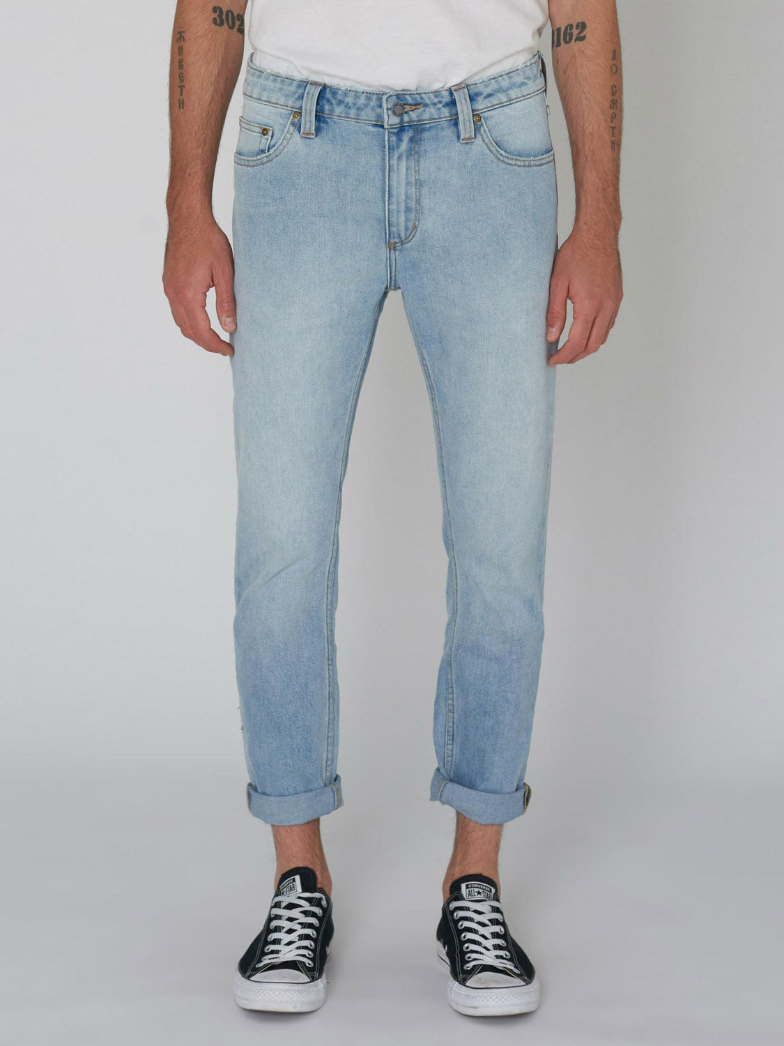 Wearing Rollas Rollies Slim Fit Jean with rolled cuffs, a white t-shirt, and black low-top sneakers, a person stands against a plain background, their tattooed forearms and wrists clearly visible.