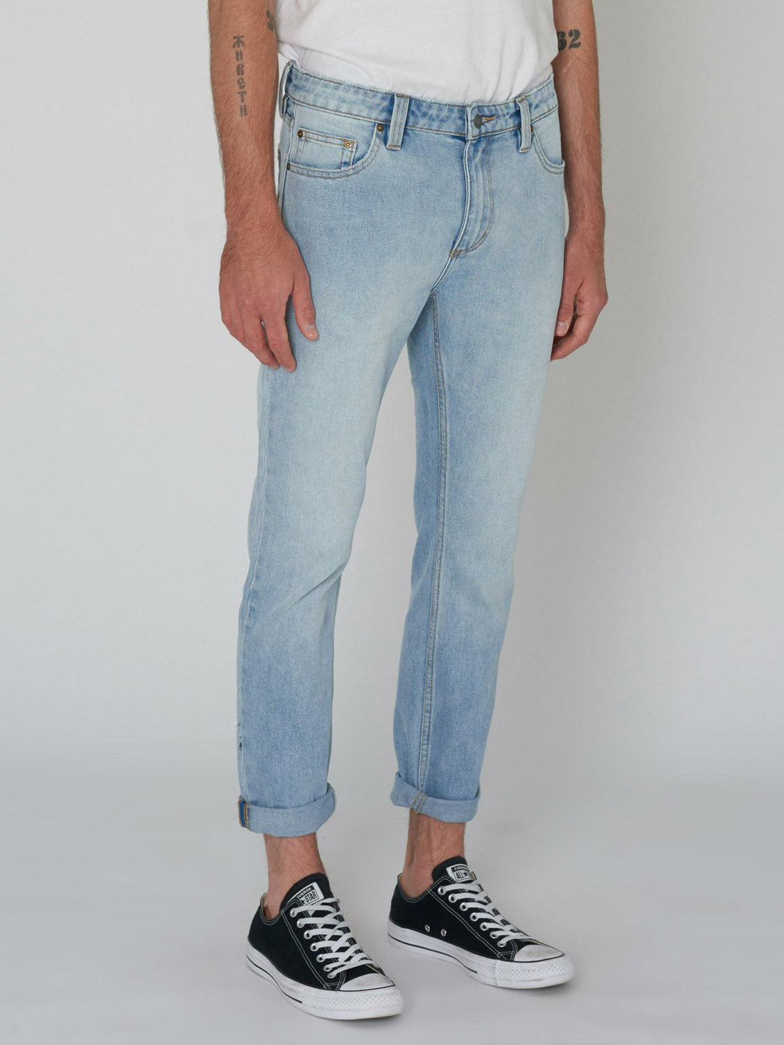 A person models Rollas Rollies Slim Fit Jean in light blue with a white t-shirt and black low-top sneakers against a white background, showing only the lower body and part of the arms.