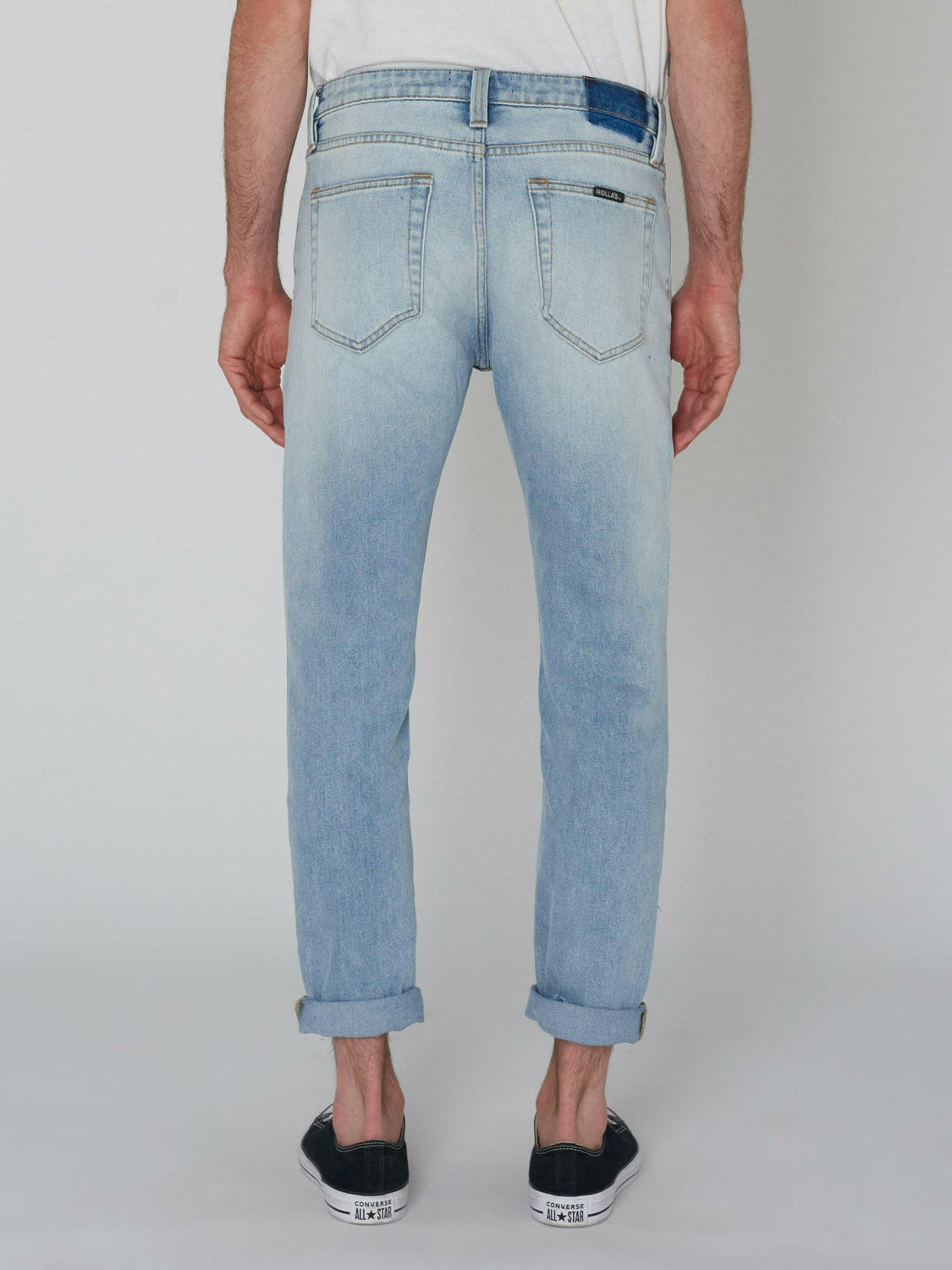 A person is seen from behind wearing Rollas Rollies Slim Fit Jean in light blue, slightly faded with rolled cuffs, paired with black slip-on shoes against a plain light background.