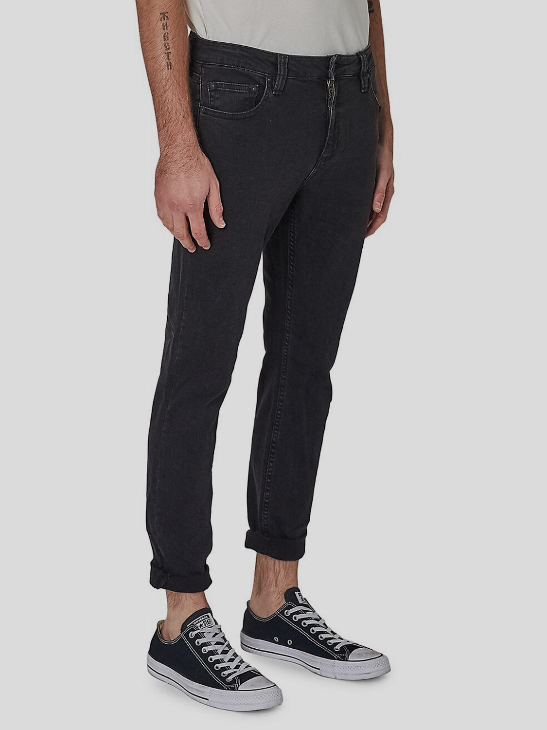 A person wearing Rollas Rollies Slim Fit Jean, a white shirt, and black low-top Converse stands on a light gray background, with the jeans cuffed at the ankles and a small tattoo visible on the left forearm.