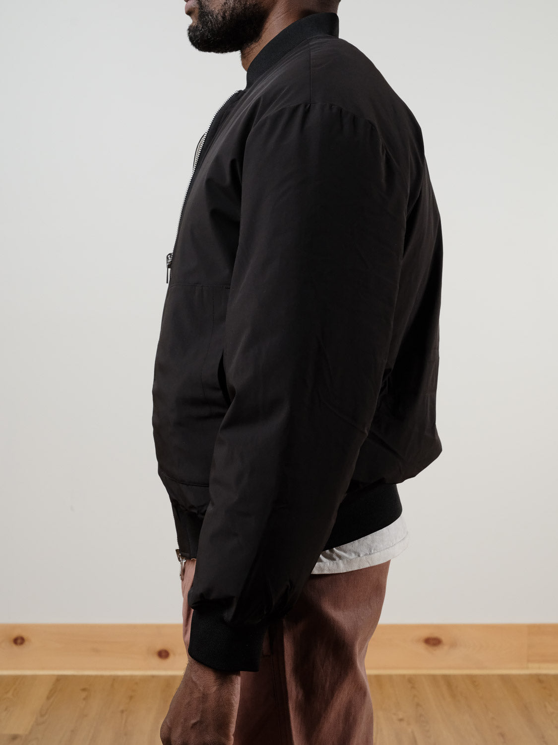 A man stands in profile wearing the !SOLID Romeo Reversible Bomber Jacket in black, paired with a white shirt and brown pants. The plain background features light-colored walls and a wooden floor.
