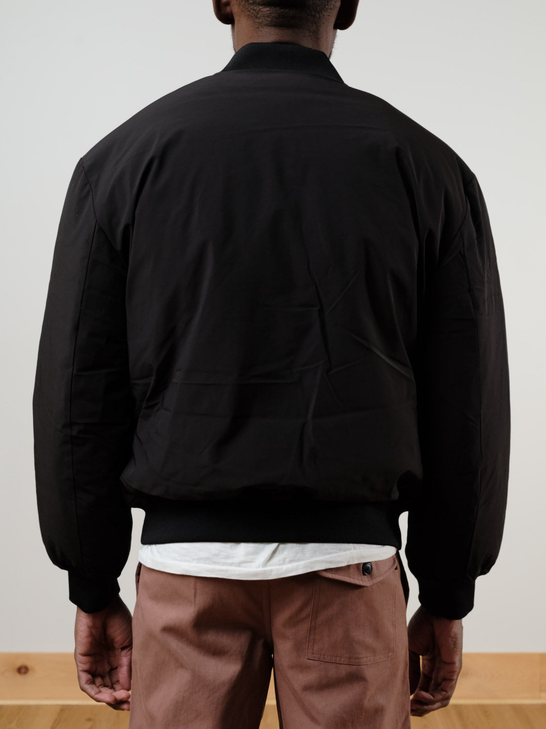 A person stands with their back to the camera, wearing the !SOLID Romeo Reversible Bomber Jacket over a white shirt and light brown pants in a minimalist indoor setting.
