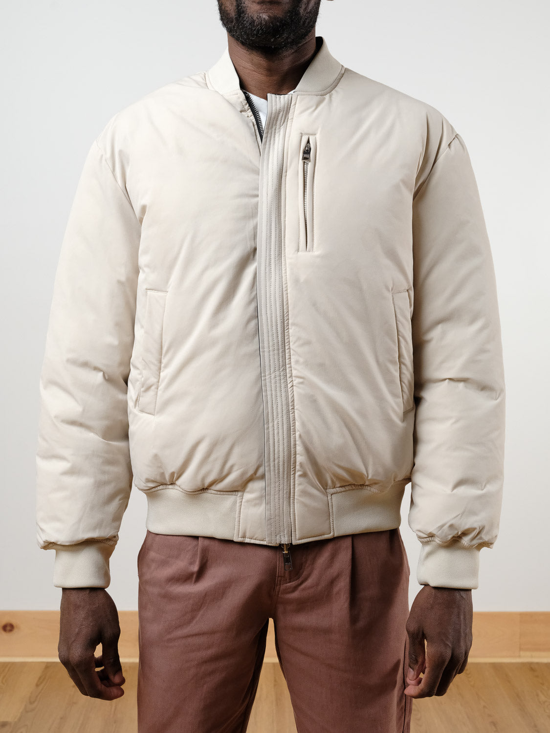 A person wears the !SOLID Romeo Reversible Bomber Jacket in cream over a white shirt and light brown pants, standing indoors on a wooden floor and facing the camera, with their head partially out of frame.