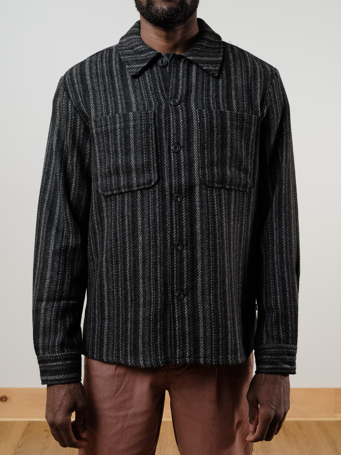 A person wearing the !SOLID Rowe Wool Blend Stripe Button-Up Shacket in grey stripes with two chest pockets and light brown pants, standing indoors against a plain background.