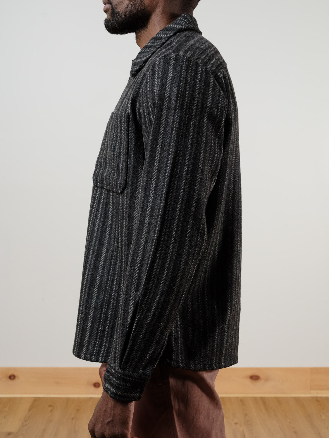 A person stands sideways in the !SOLID Rowe Wool Blend Stripe Button-Up Shacket—grey with thin, light vertical stripes and a chest pocket—paired with light brown pants, against a plain light wall and wooden floor.