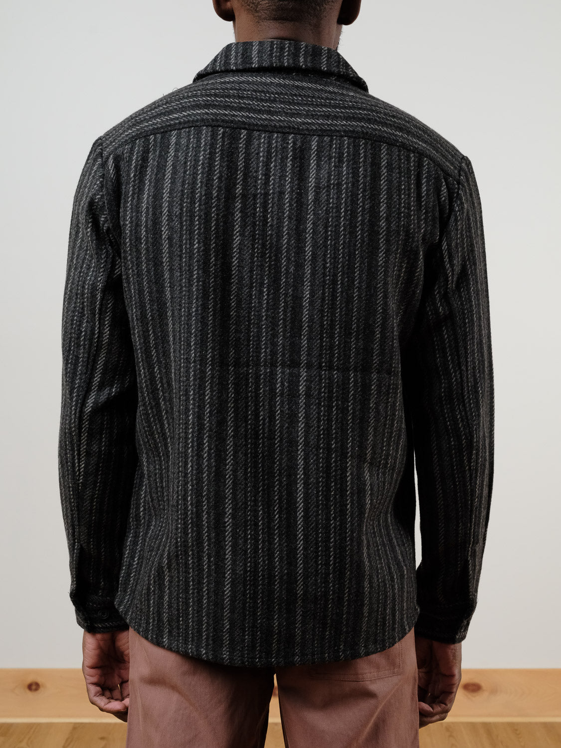 A person stands with their back to the camera, wearing the !SOLID Rowe Wool Blend Stripe Button-Up Shacket in dark grey and light brown pants, set against a plain light background and wooden floor.