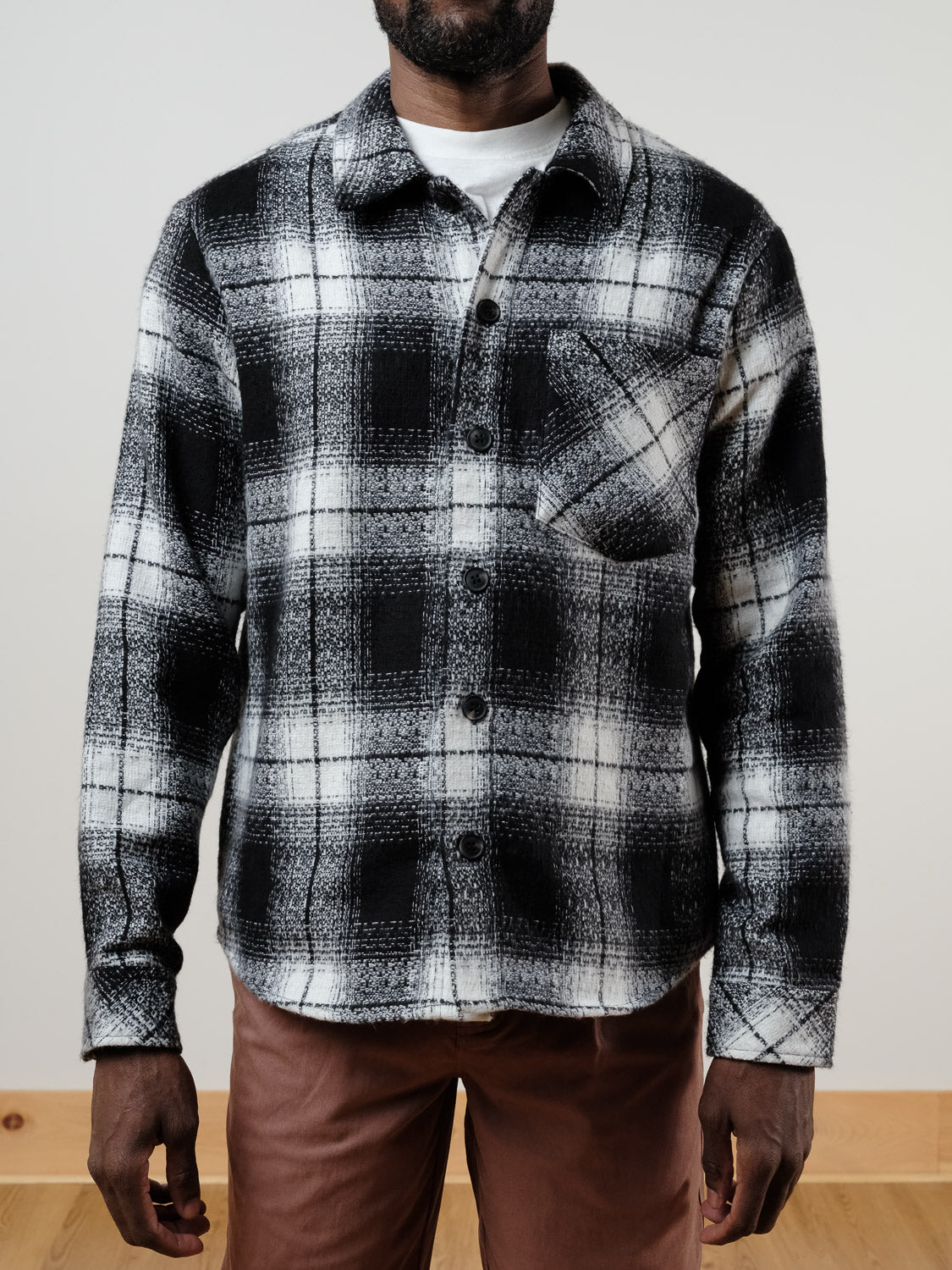 A person wears the !SOLID Ruhi Plaid Button Down Shacket over a white t-shirt and relaxed-fit brown pants, standing indoors against a plain background.