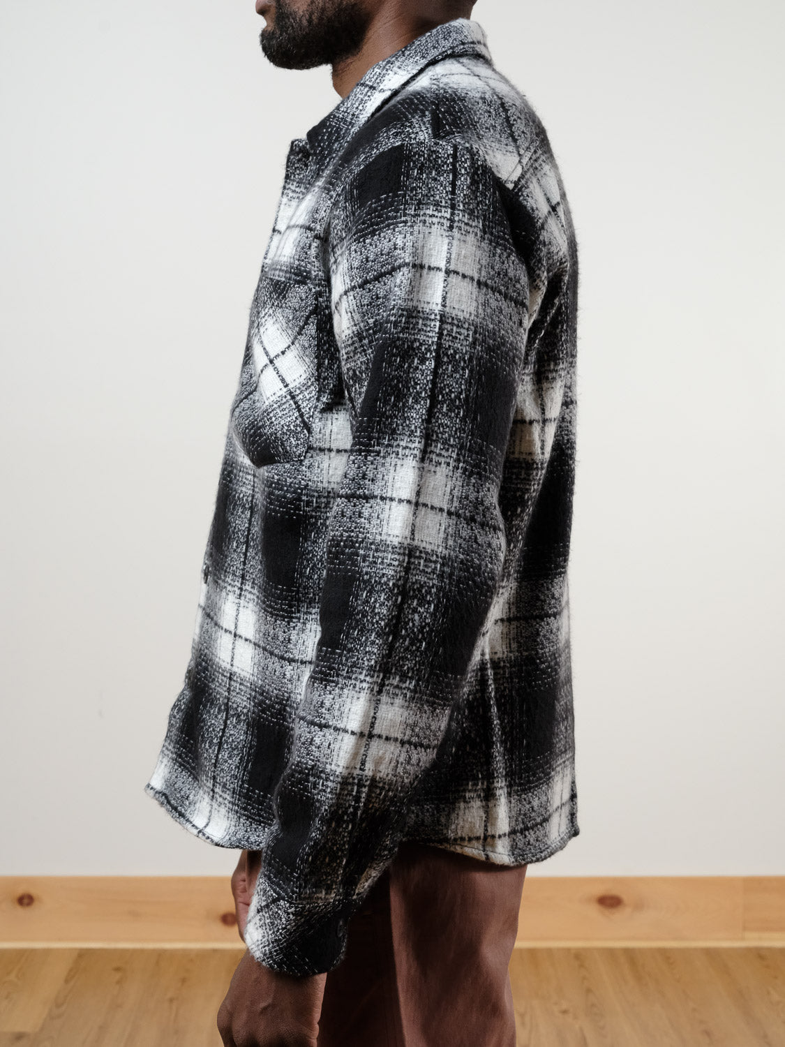 A person wearing the !SOLID Ruhi Plaid Button Down Shacket in black and white with brown pants stands sideways in a room with light-colored walls and wooden flooring, their face not visible.