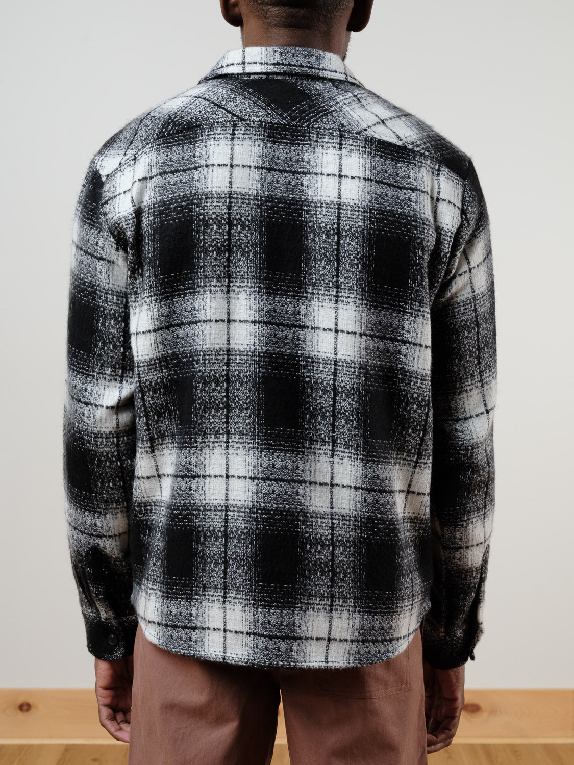 A person faces away, wearing the !SOLID Ruhi Plaid Button Down Shacket in black and white with long sleeves, paired with relaxed-fit light brown pants, standing against a plain light wall and wooden floor.