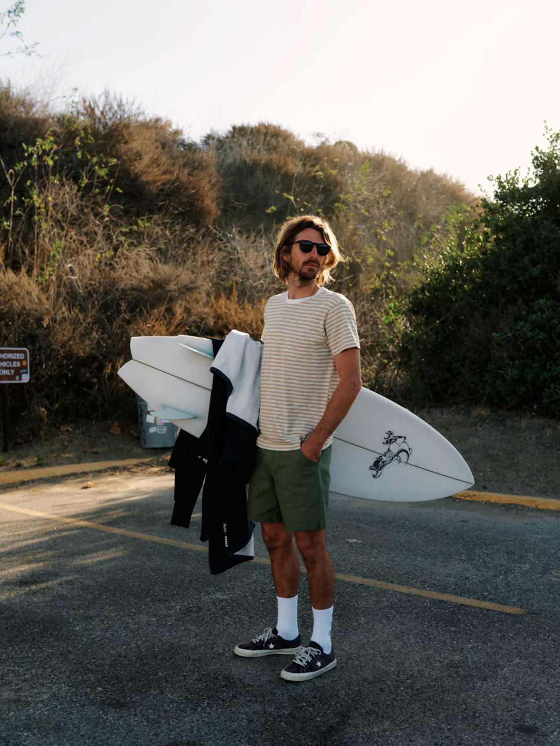 A man with long hair and sunglasses stands on a paved road, holding a surfboard and black wetsuit. He wears the Katin Rural Pocket Tee, green shorts, white socks, and sneakers. Bushes and a sign are visible in the background.