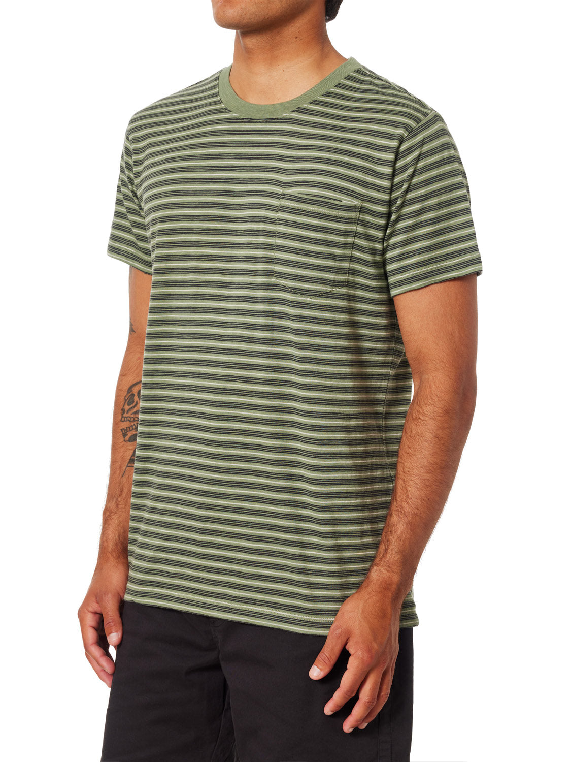 Sporting Katins Rural Pocket Tee in premium cotton with green and black stripes, a small chest pocket, and short sleeves, the individual pairs it with black pants. The round-neck tee reveals a tattoo on their left forearm against a plain white backdrop.