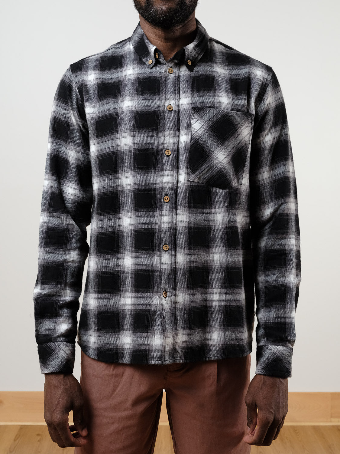 A person wearing the !SOLID Rye Organic Cotton Plaid Button-Down Shirt in black and white with a chest pocket, paired with brown pants, stands indoors against a plain light background.