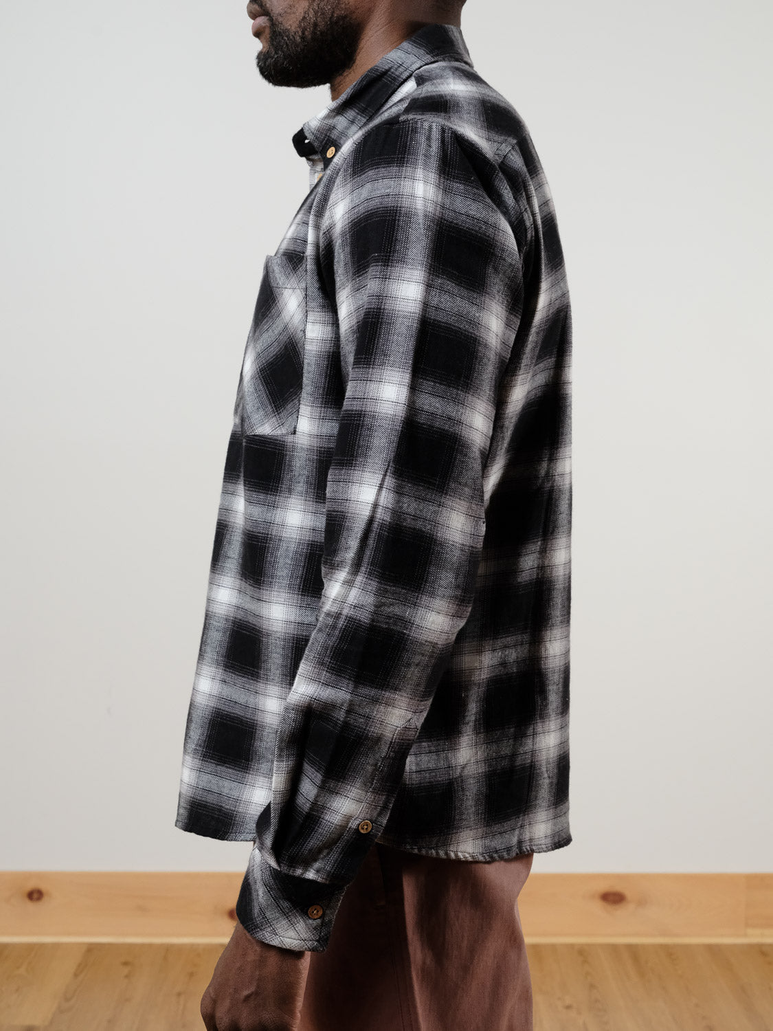 A person stands sideways wearing the !SOLID Rye Organic Cotton Plaid Button-Down Shirt in black and white and brown pants, against a light-colored wall with a wooden floor in the background.
