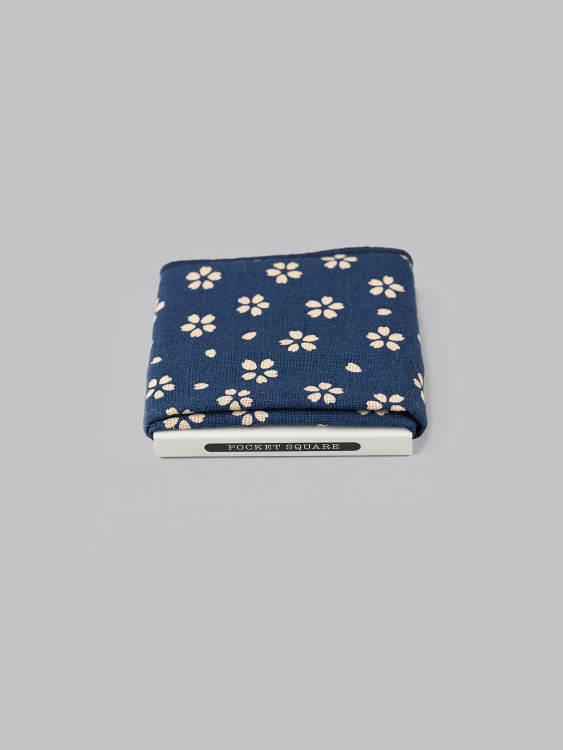 The Sakura Pocket Square by ID Menswear is a navy blue cotton accessory with a white floral pattern, handmade in Brooklyn and displayed on a light gray background.
