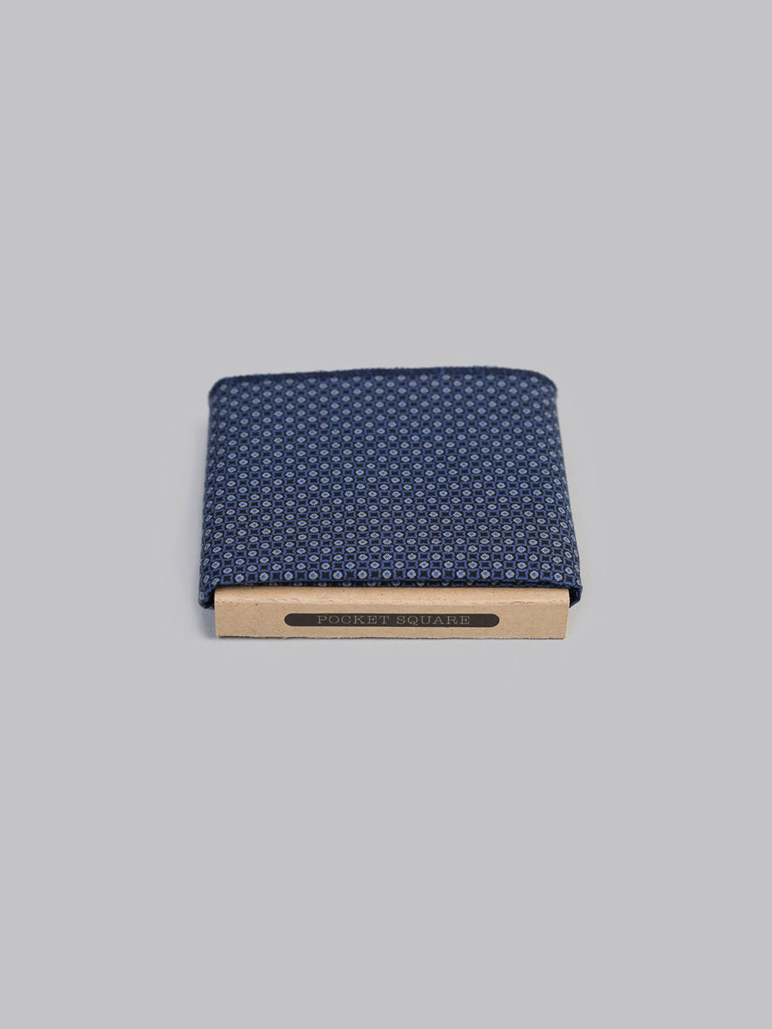 The Sakuru Pocket Square by ID Menswear features a neatly folded blue Japanese print with a small geometric pattern, handmade in Brooklyn. It is partially tucked into a brown POCKET SQUARE holder and displayed against a plain gray background.