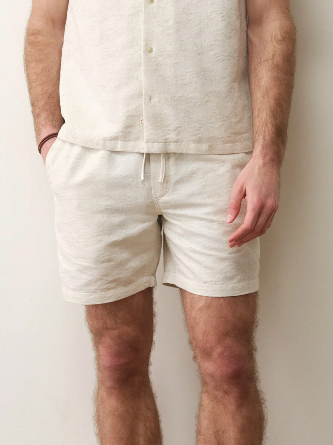A person stands against a plain wall, wearing a light beige short-sleeve shirt and Marine Layer Saturday Beach Shorts 6" with a subtle leaf jacquard texture. One hand is in the stash pocket; their face is not visible.