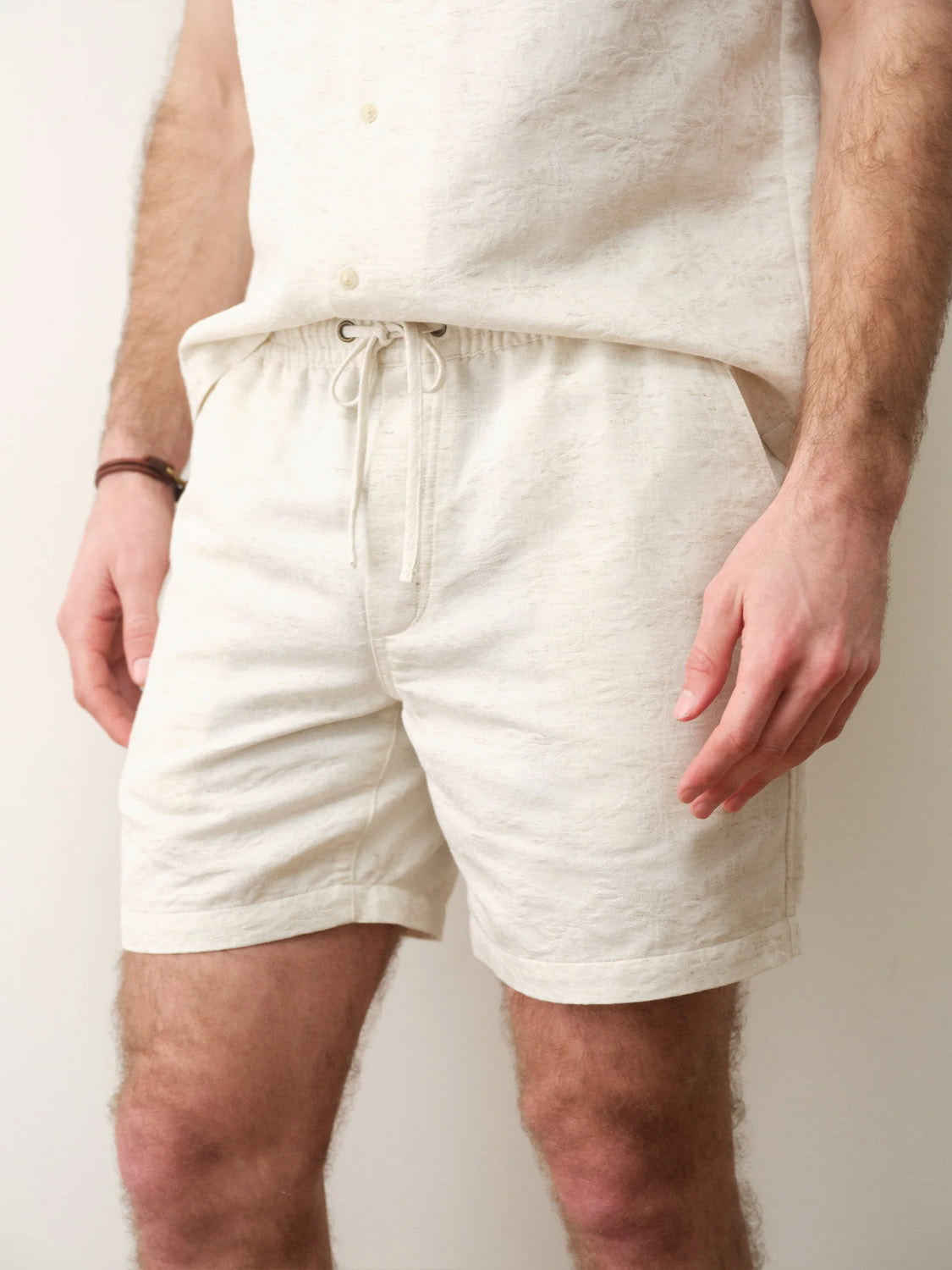 A person models Marine Layer's Saturday Beach Short 6" in a matching off-white set, standing against a plain light background. The casual, comfortable outfit is showcased with the person's head out of frame.