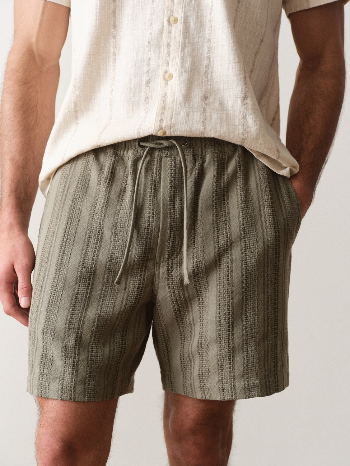 A person wears Marine Layer's Saturday Dobby Short 6" in olive green stripes, paired with an off-white, short-sleeve button-up shirt. Their hands rest by their sides; the image highlights their lower torso and upper legs.