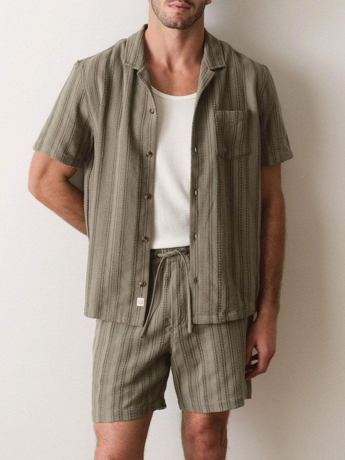 A man stands against a plain wall wearing a light olive green Marine Layer Saturday Dobby Short 6" and matching short-sleeved button-up shirt in textured cotton over a white tank top. His face is partially out of frame.