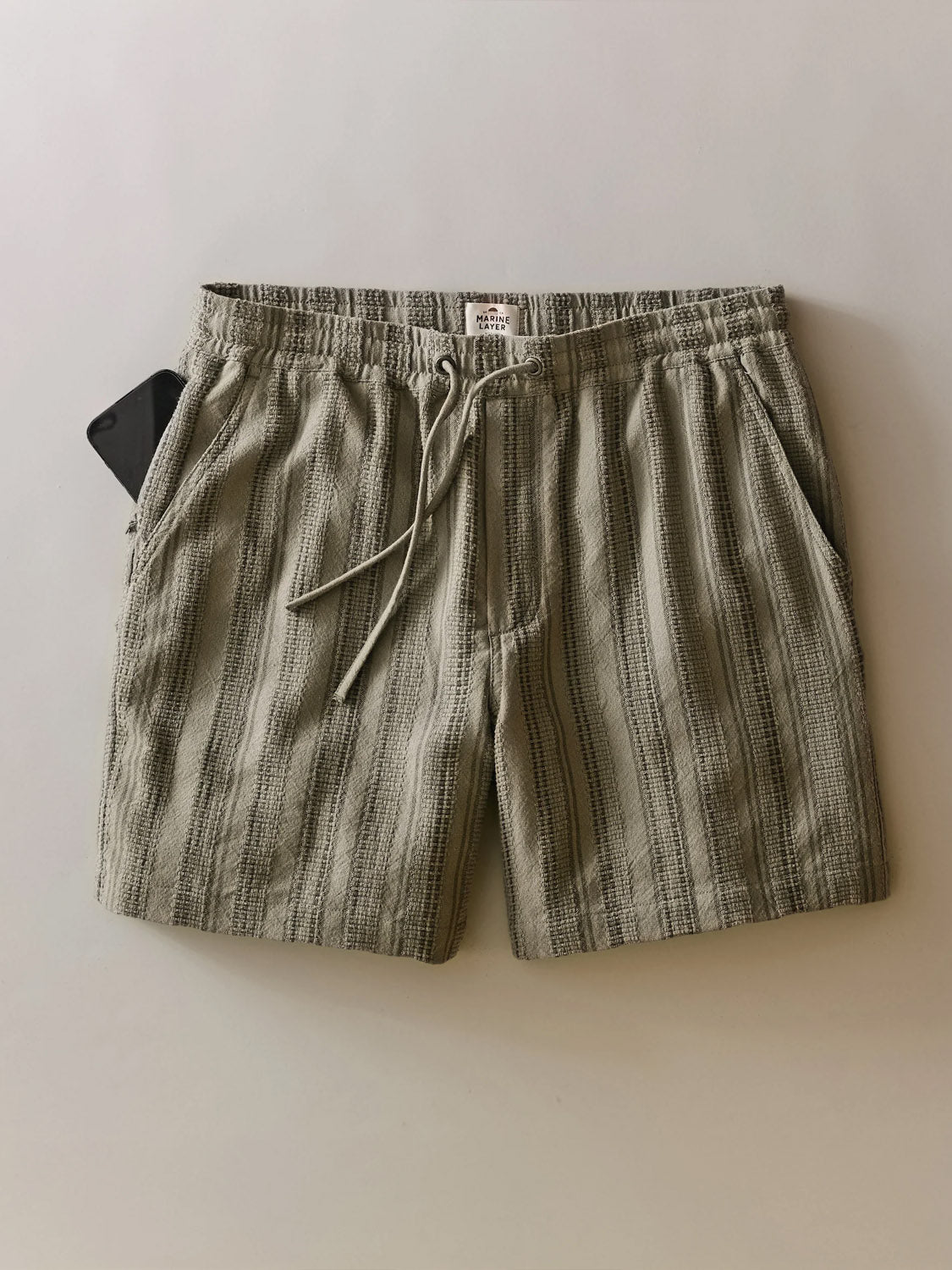 The Marine Layer Saturday Dobby Short 6" in green and beige stripes features a 100% cotton dobby weave, two side pockets, a waistband label, and is shown laid flat with a black smartphone partially in the left pocket.
