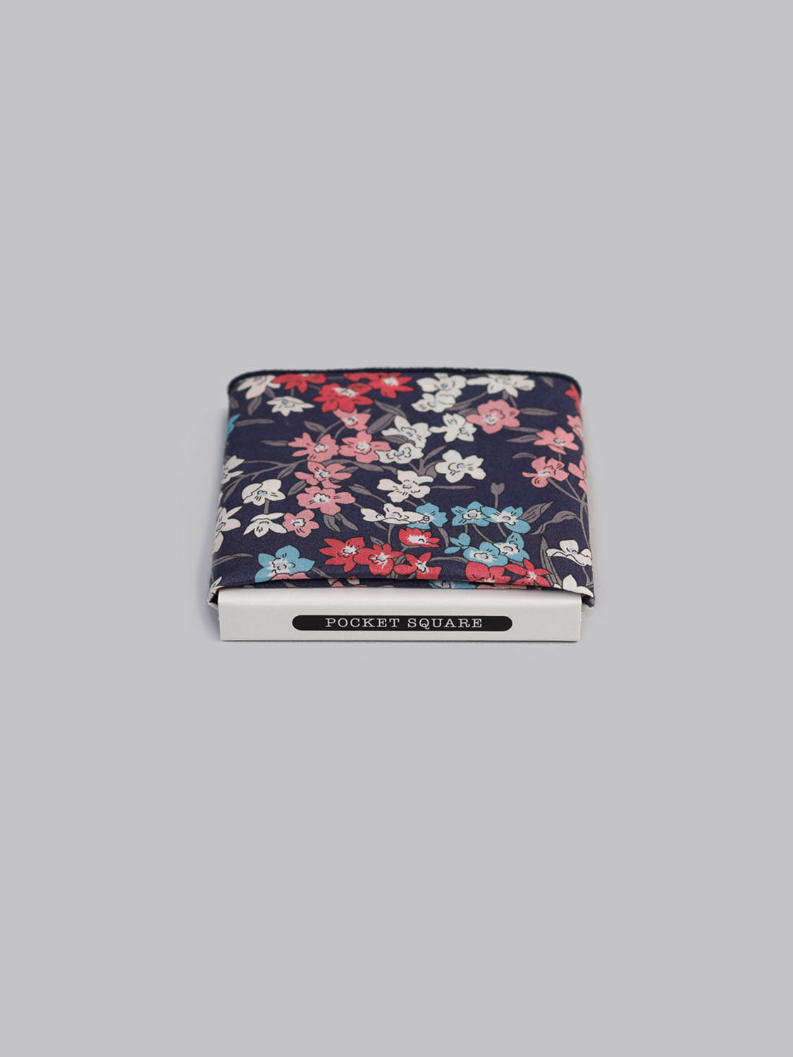 A Liberty™ Sea Bloom Pocket Square by ID Menswear, handmade in Brooklyn from fine Tana Lawn cotton, features a navy blue background with red, pink, white, and blue floral print. Displayed folded in a white POCKET SQUARE box on a light gray backdrop.
