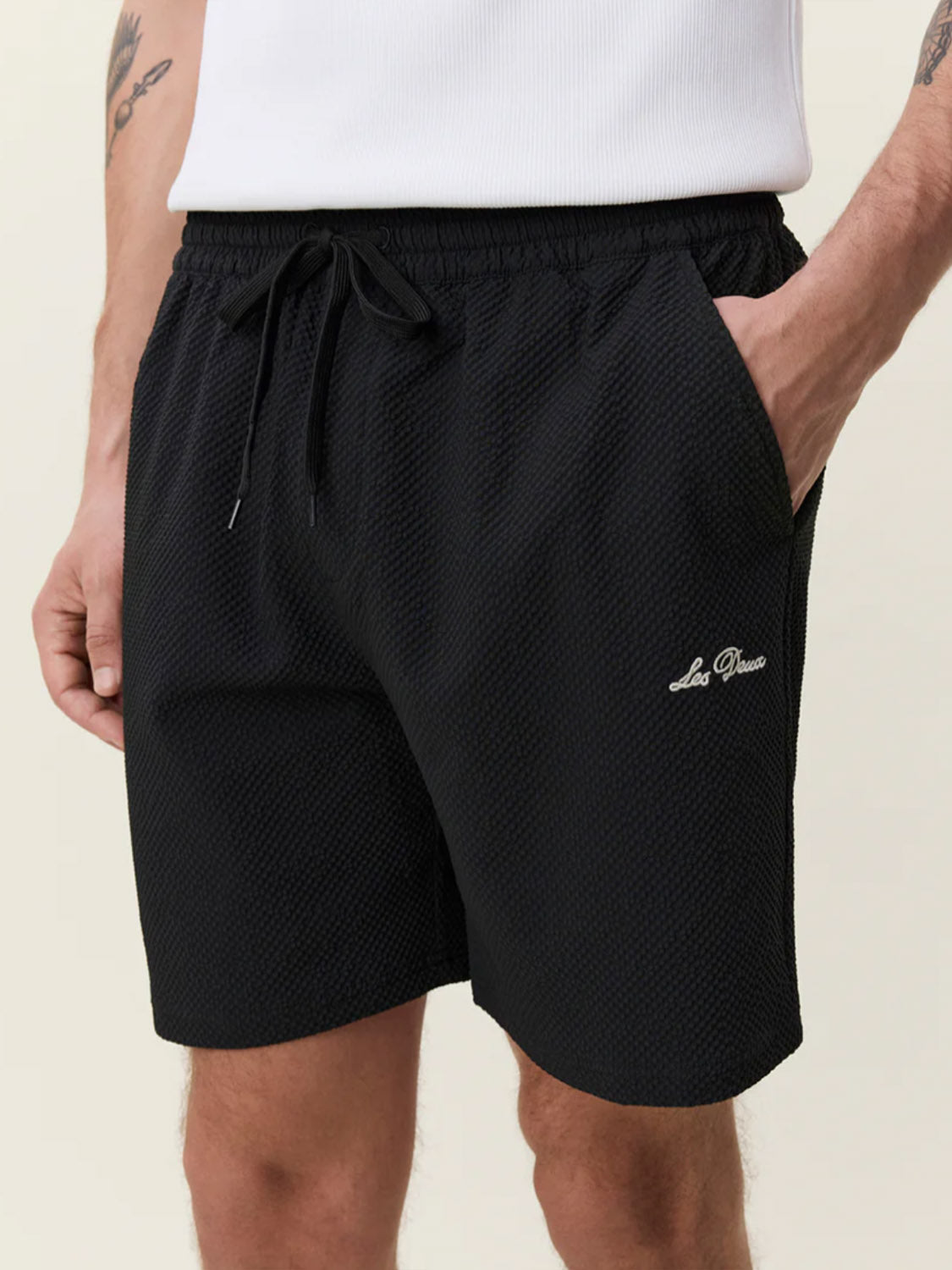 A person wears Les Deux Seersuckers Swim Shorts—black, seersucker textured, with a drawstring and small white logo on the left leg—paired with a white sleeveless top and one hand in their shorts pocket.