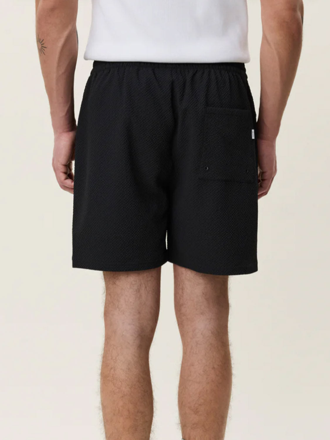 A person is seen from behind wearing Les Deux Seersuckers Swim Shorts in black, featuring a seersucker texture, elastic waistband, and right-side back pocket. They are also wearing a white shirt against a neutral background.