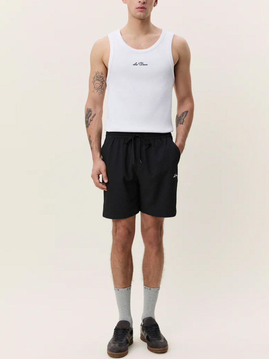 A person wearing a white tank top, Les Deux Seersuckers Swim Shorts in black, gray socks, and black shoes stands against a plain light background. Tattoos are visible on both arms.