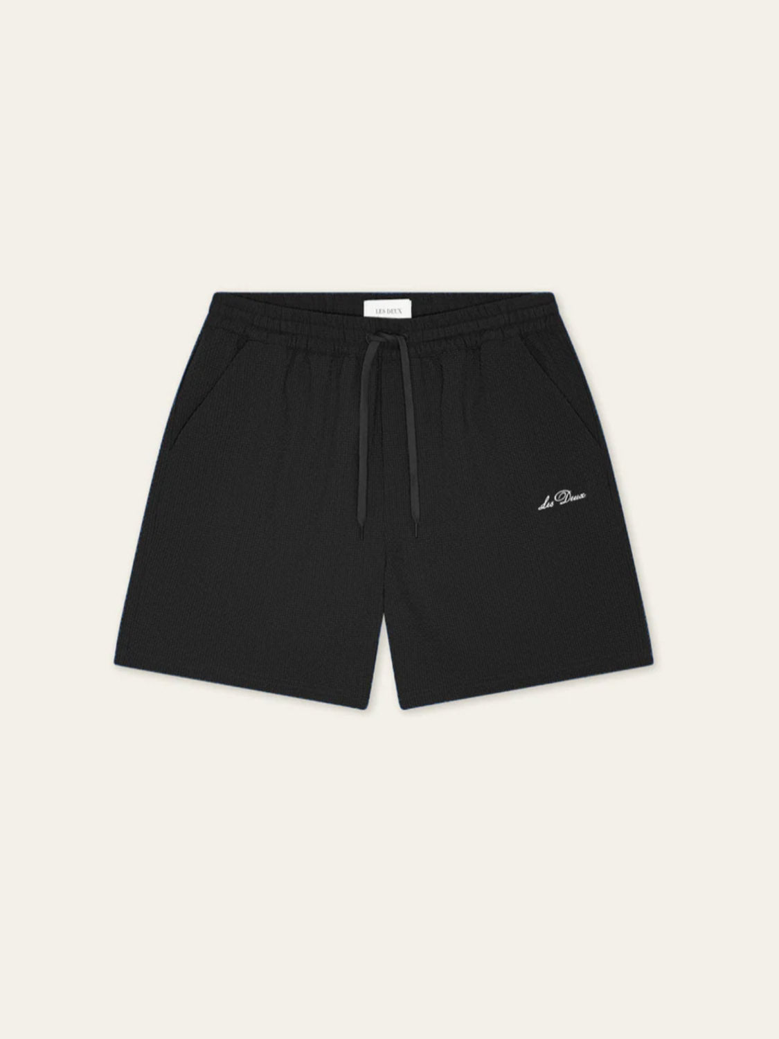 Les Deux Seersuckers Swim Shorts in black feature side pockets, a small white logo on the left leg, and are made from recycled polyester with a subtle seersucker texture, shown against a plain light background.
