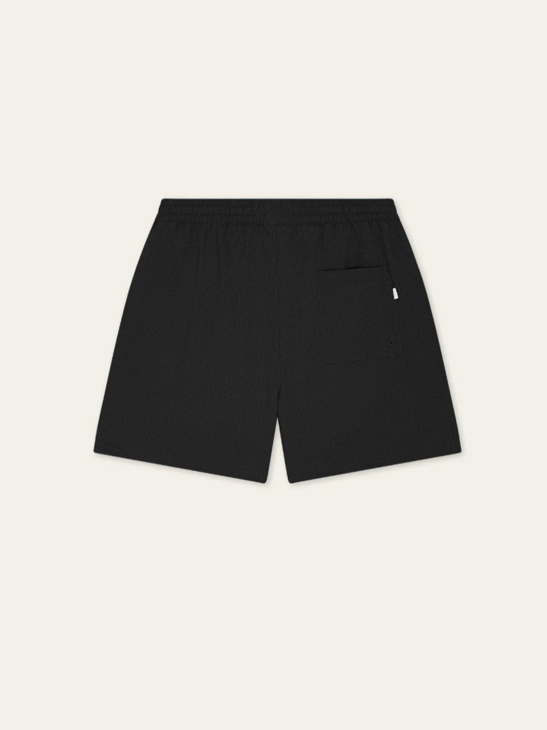 Les Deux Seersuckers Swim Shorts in black, shown from the back with an elastic waistband and a single right patch pocket. Made from recycled polyester with a subtle seersucker texture. Off-white background.