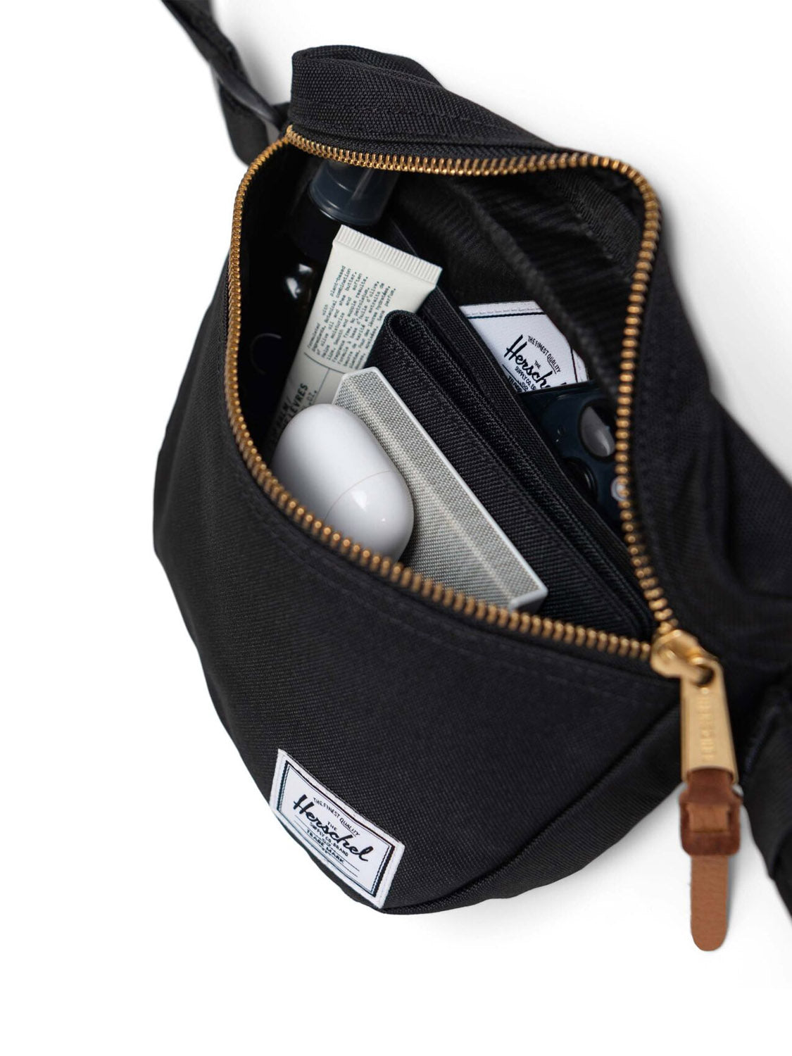 The Herschel Settlement Hip Pack in black recycled fabric, featuring a brown leather zipper pull, is open to reveal a tube of cream, wallet, white oval item, and other small essentials—ideal for hands-free journeys.