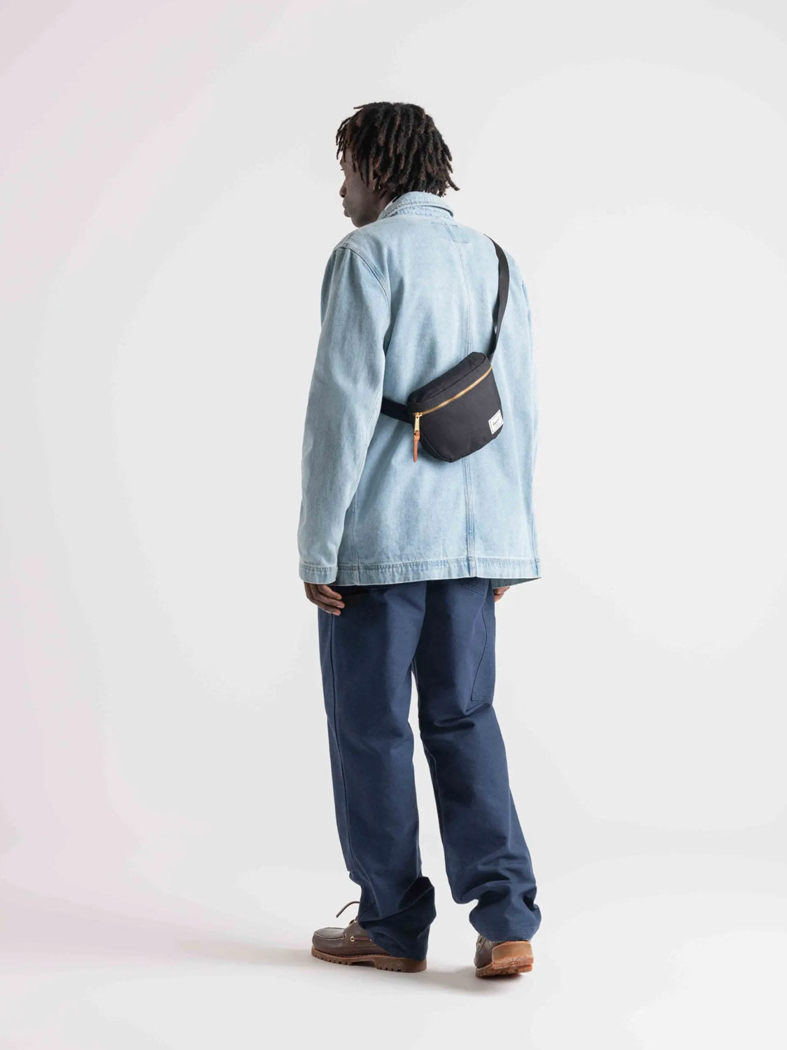 A person with short dreadlocks faces away, wearing a light blue denim jacket, dark blue pants, brown shoes, and the Herschel Settlement Hip Pack - 2L (black, recycled polyester) for hands-free journeys against a plain white background.