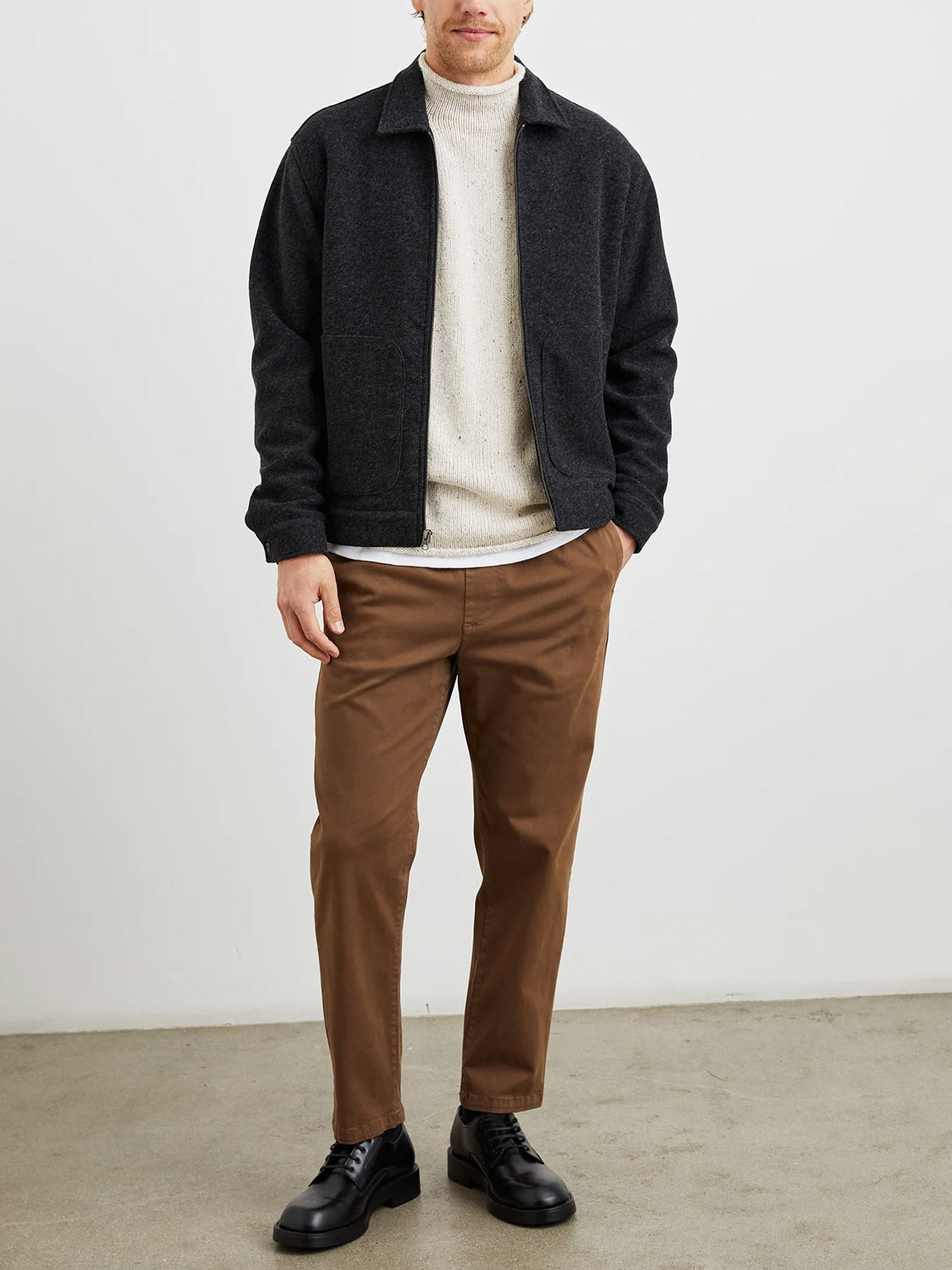 A man stands indoors against a plain wall, wearing the Rails Shael Jacket in dark gray over a cream sweater, with brown pants and black shoes. His hands are in his pockets and his face is partially visible.