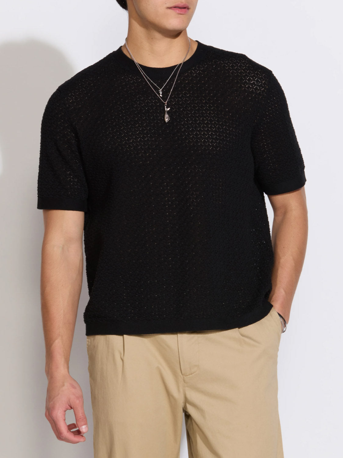 Someone stands against a plain white background, their face partially out of frame, wearing the Civil Society Shafa Boxy Fit Sweater Knit Tee in black, beige pants, and layered silver necklaces.