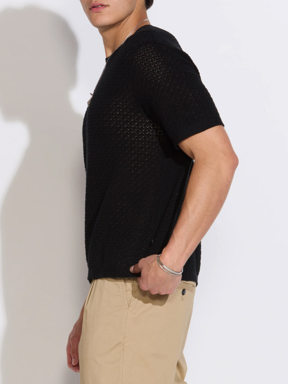 A person stands sideways against a white background wearing the Civil Society Shafa Boxy Fit Sweater Knit Tee in textured black and beige pants. Their face is partially out of frame, and they accessorize with a silver bracelet.