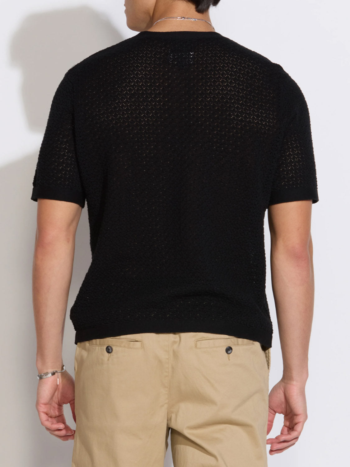Shown from the back, a person with short dark hair wears the Civil Society Shafa Boxy Fit Sweater Knit Tee in black and light beige pants. Textured mesh knit adds style. Accessories include a bracelet and necklace; background is plain and light-colored.