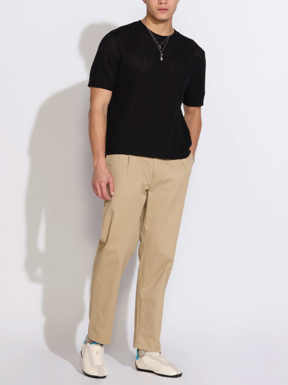 A man stands against a plain white background with one hand in his pocket, wearing the Civil Society Shafa Boxy Fit Sweater Knit Tee in black, paired with beige pants and white sneakers.