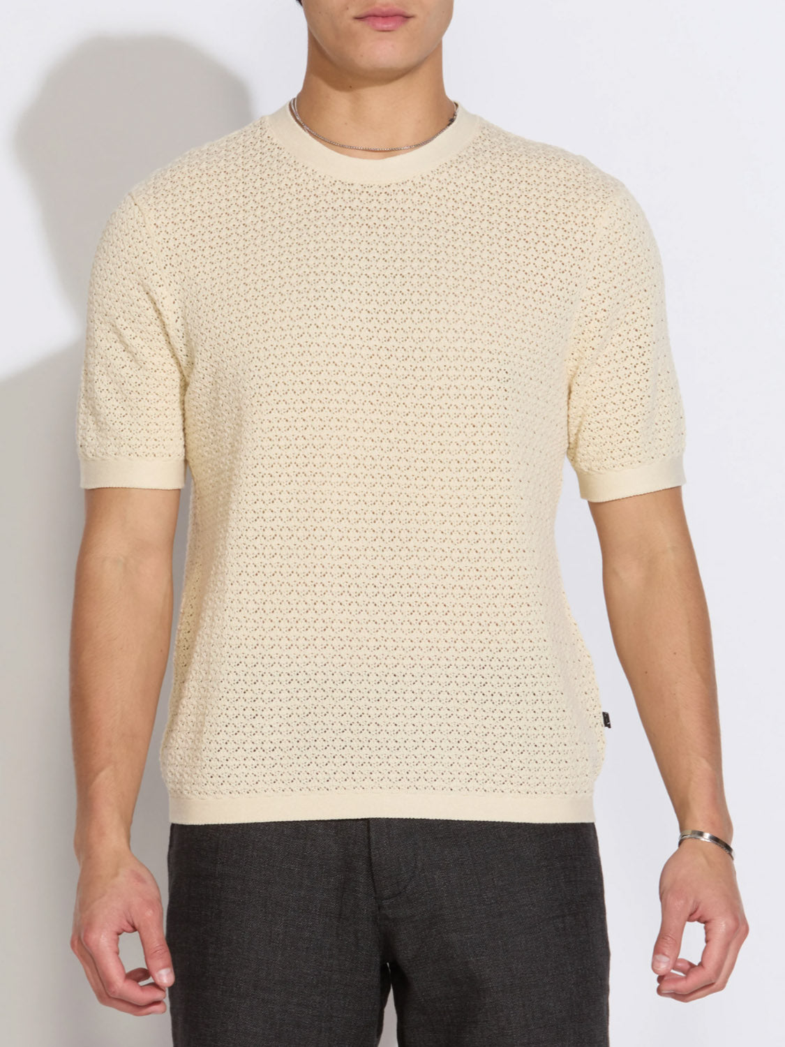 A person wears the Civil Society Shafa Boxy Fit Sweater Knit Tee, a cream short-sleeve top with subtle mesh knitting, paired with dark trousers and standing against a plain light background.