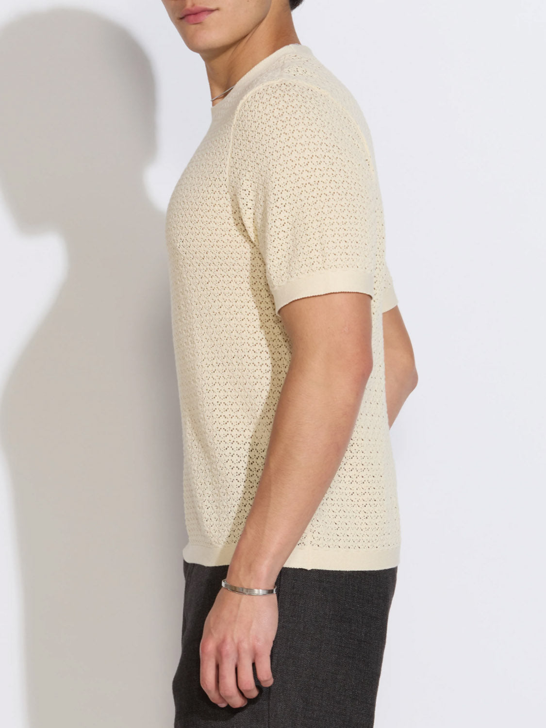 A man stands sideways against a white background, wearing the Civil Society Shafa Boxy Fit Sweater Knit Tee in light beige with mesh knitting and dark pants. Only his upper half is visible, and he sports a simple bracelet on his wrist.
