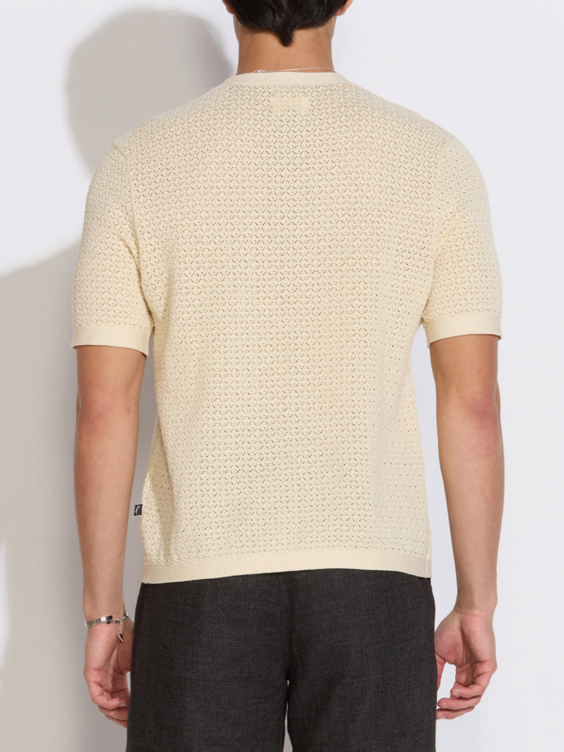 A person is seen from behind in a Civil Society Shafa Boxy Fit Sweater Knit Tee, cream-colored with a textured finish, paired with dark pants and standing against a plain white background.