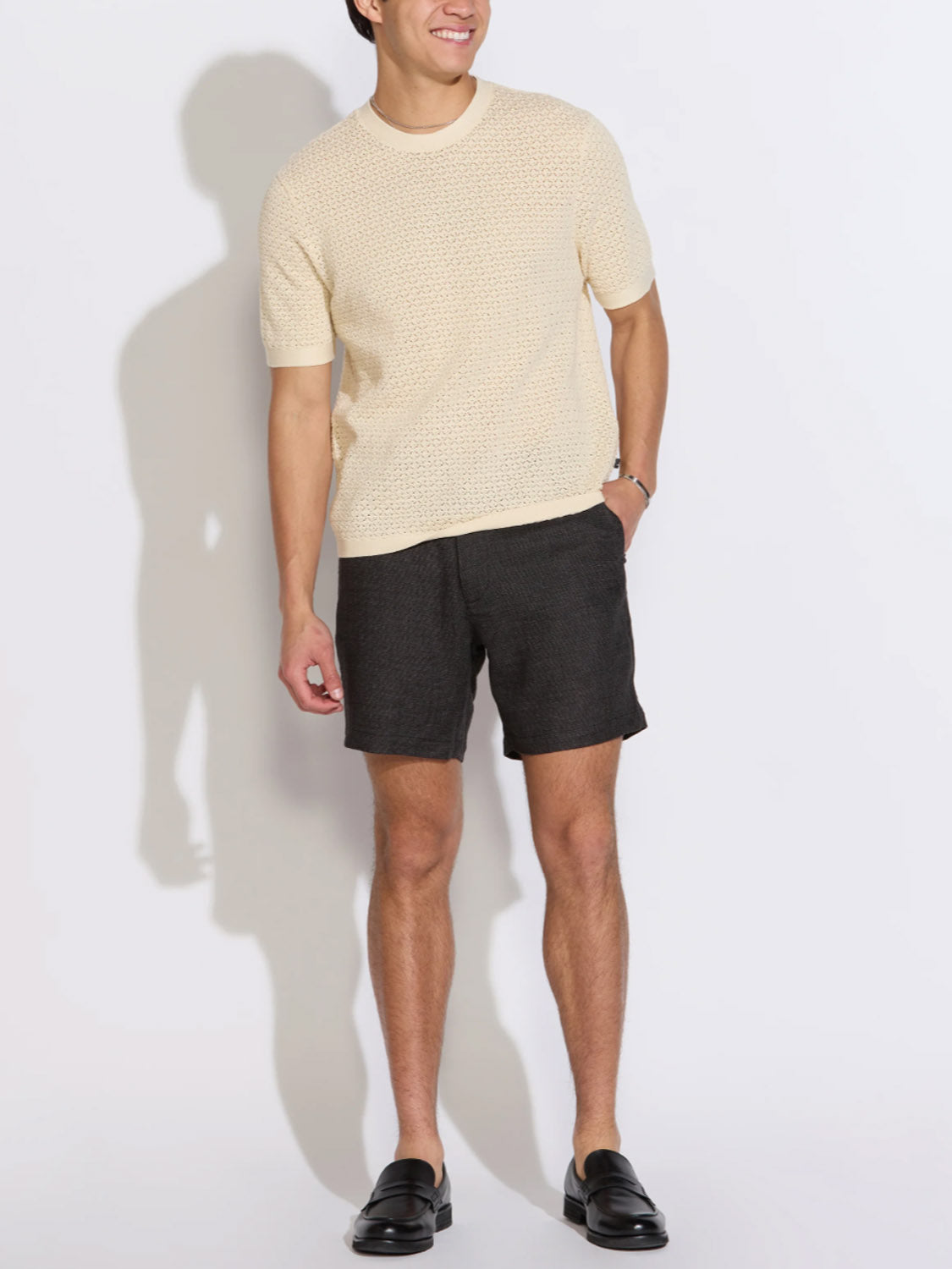 A person stands against a white background, smiling with one hand in their pocket, wearing the Civil Society Shafa Boxy Fit Sweater Knit Tee, dark gray shorts, and black loafers.