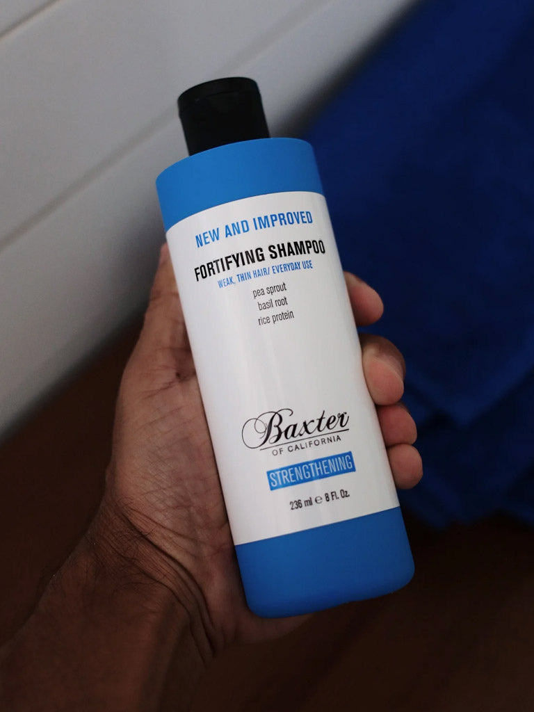 A hand holds a blue and white bottle of Baxter Fortifying Shampoo, a thickening, strengthening shampoo for everyday use, featuring a new and improved formula and promises of full, strong hair on the label.