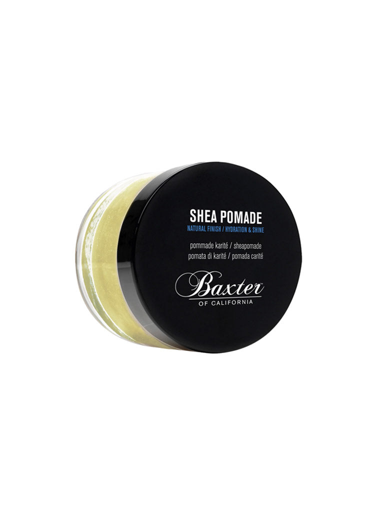 A round container of Baxter Shea Pomade Natural Finish and Shine features a black lid, white and blue text, and pale yellow pomade inside. The label notes its moisturizing hair balm formula for hydration, shine, and a natural finish.