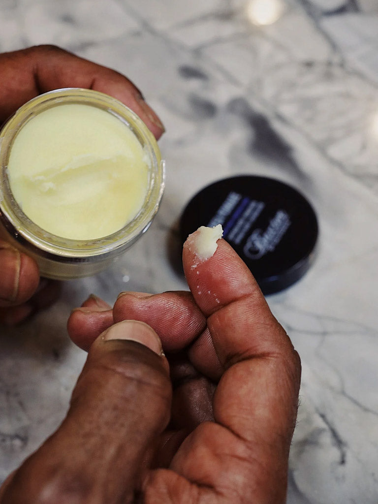 A person scoops a small amount of Baxter Shea Pomade Natural Finish and Shine from its jar onto their fingertip, with the lid resting on a marble surface in the background.