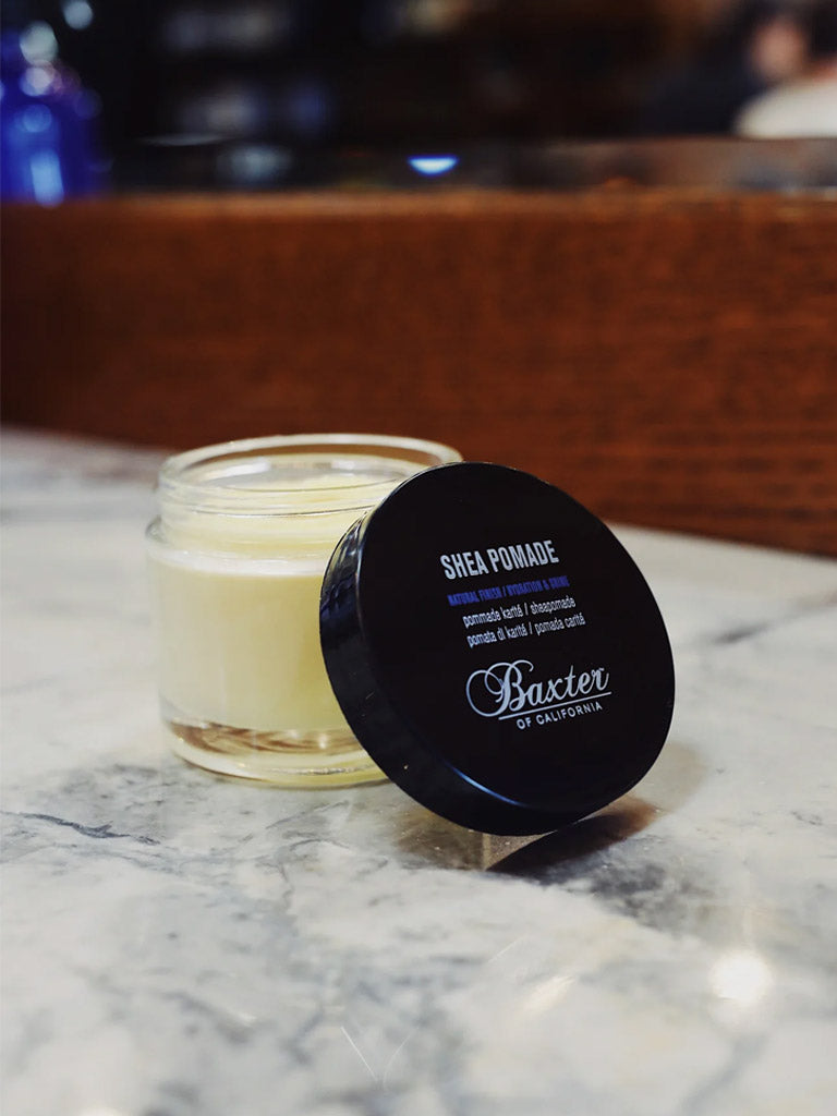 An open jar of Baxter Shea Pomade Natural Finish and Shine sits on a marble countertop, its black lid nearby. The creamy balm is ideal for textured hair. A wooden box and a softly blurred background complete the scene.