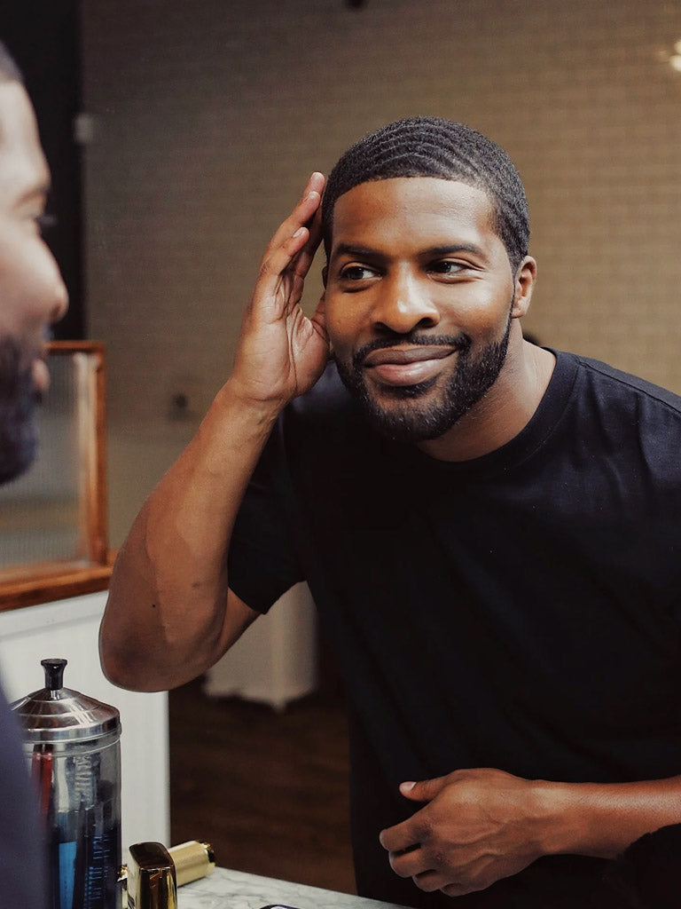 A man with textured hair and a neatly trimmed beard smiles at his reflection, touching his head after applying Baxter Shea Pomade Natural Finish and Shine. He wears a black t-shirt and stands in a well-lit room.