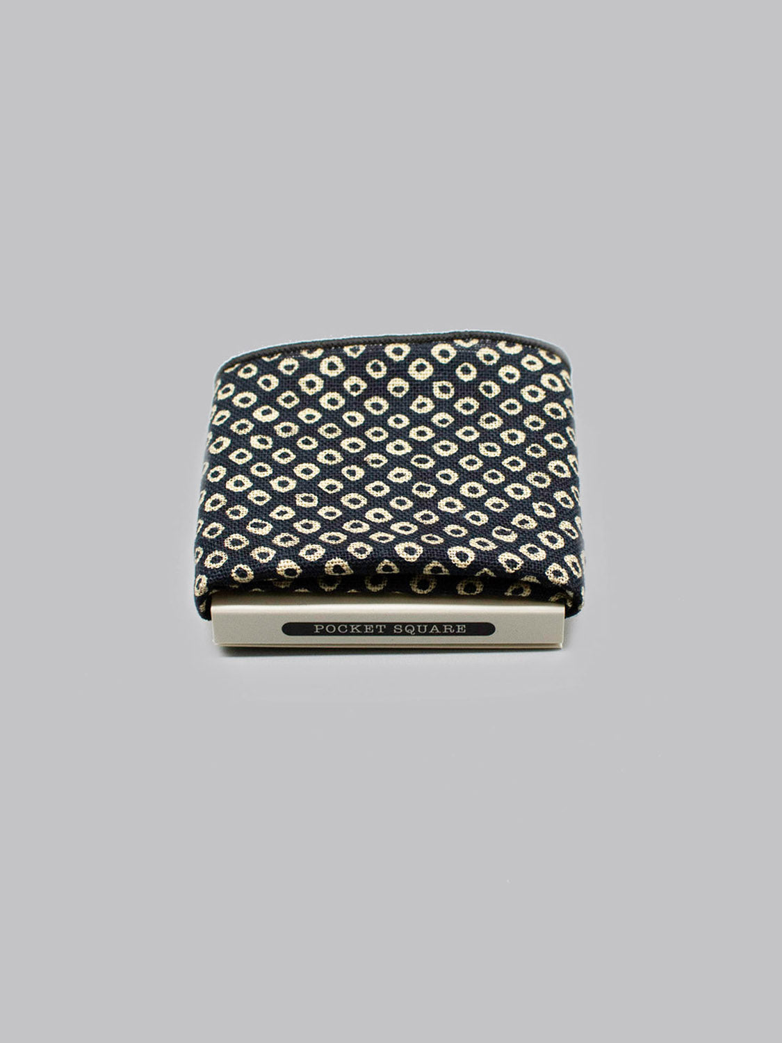 The Shibori Pocket Square by ID Menswear, featuring a black and beige geometric Japanese print, is partially tucked into a silver holder labeled POCKET SQUARE against a plain gray background.